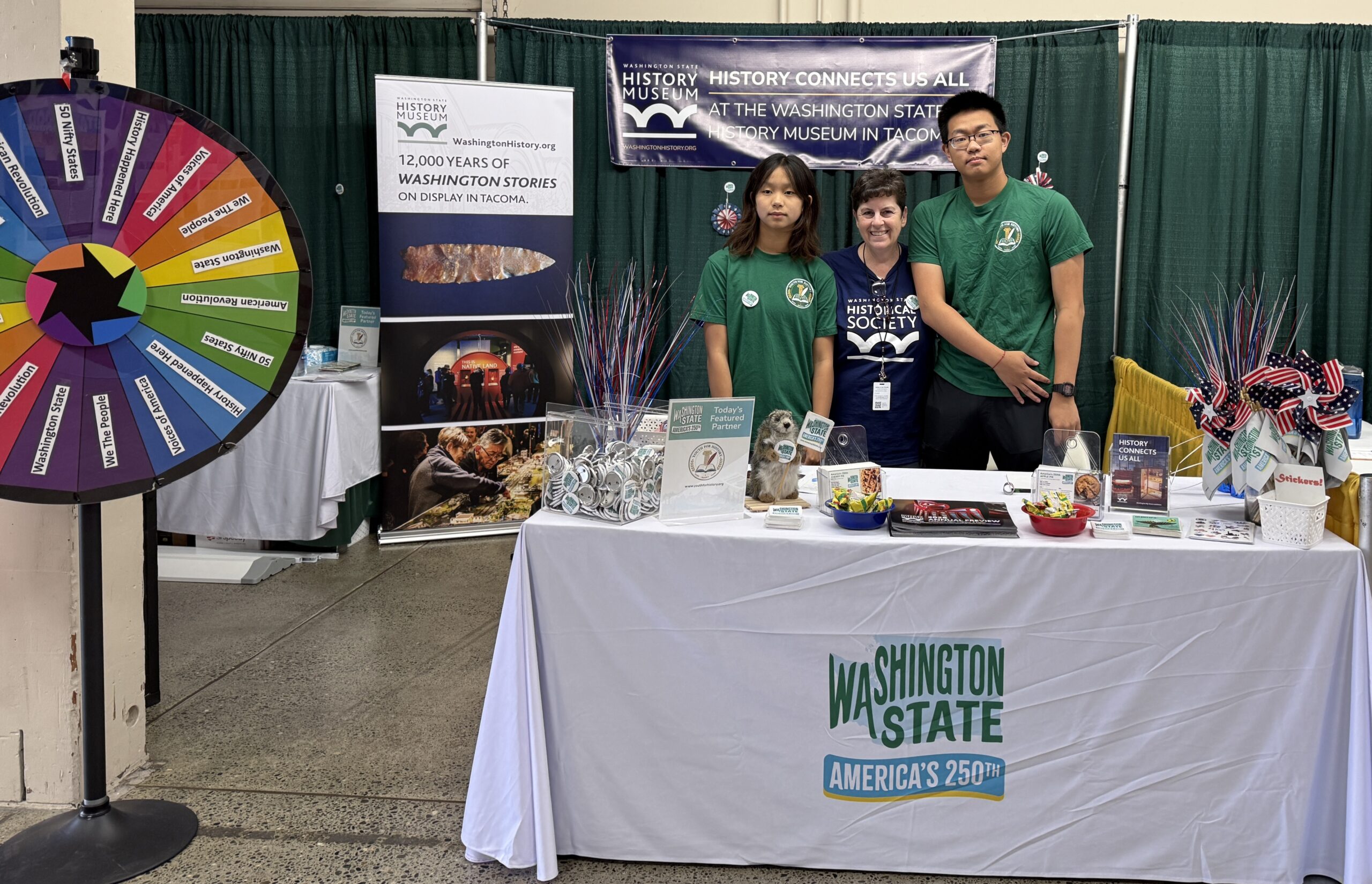 Youth volunteers and a Washington State Historical Society representative behind the history booth, with an activity wheel and America’s 250th materials.